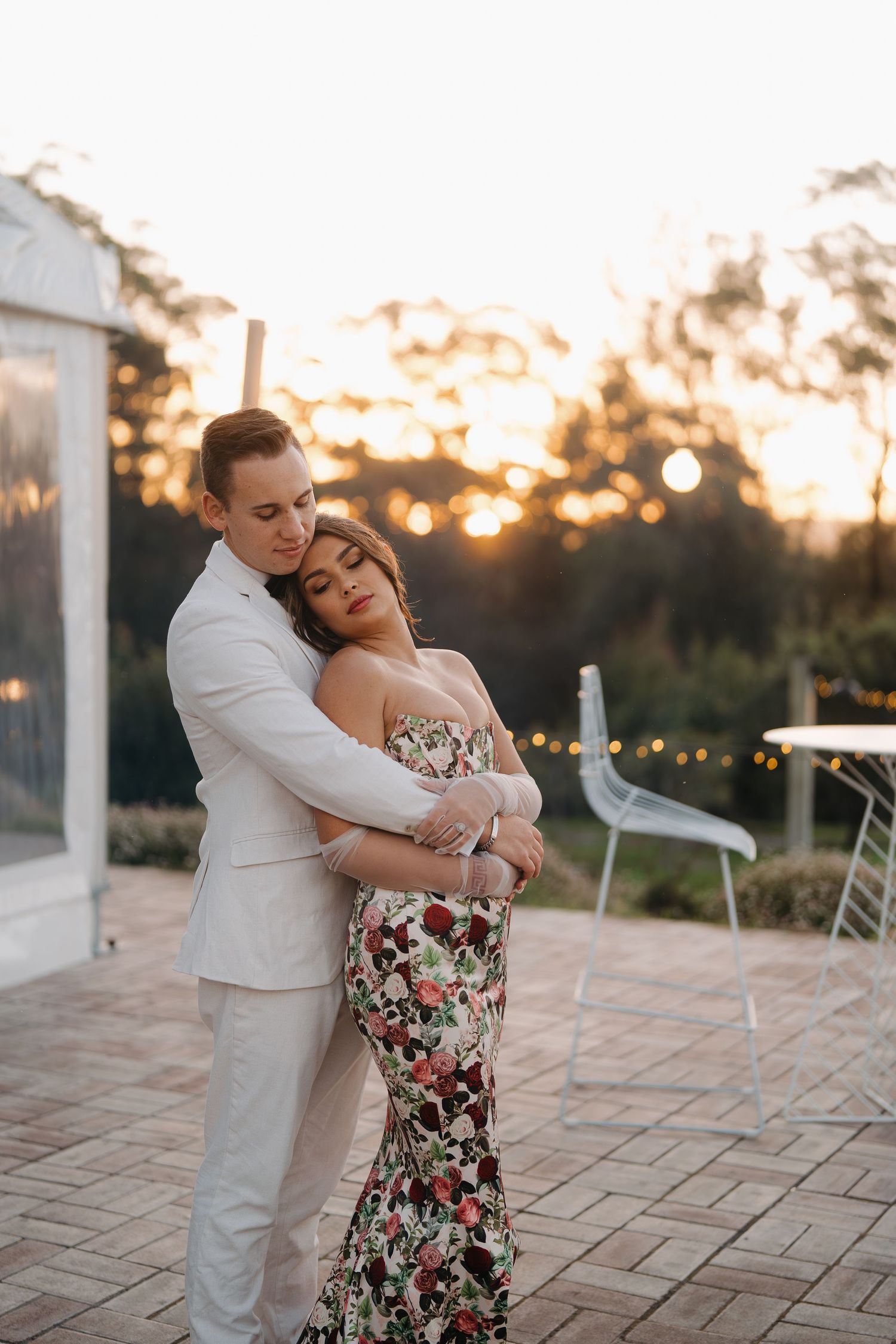 Couple embracing at sunset, man in white suit hugging woman in floral gown, romantic outdoor evening setting.