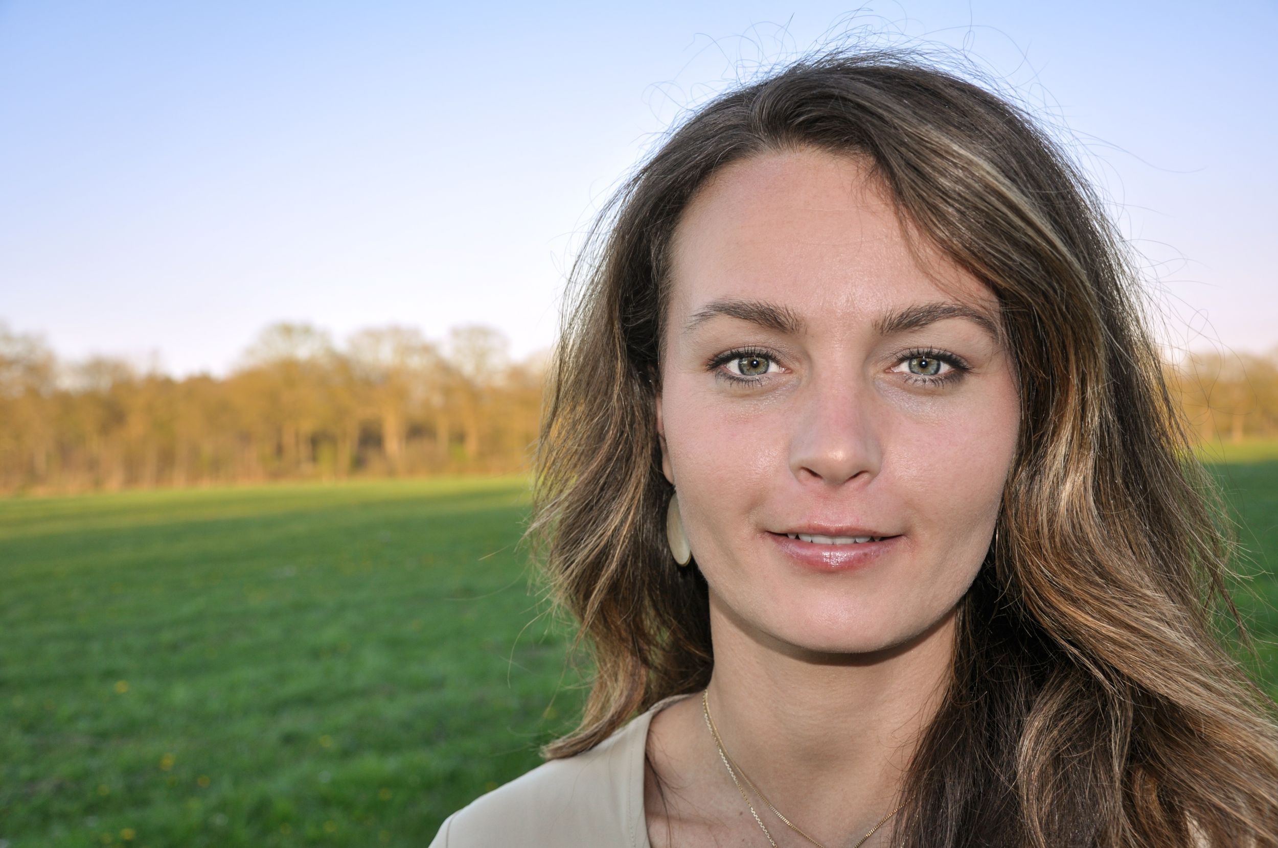 Young woman with brown hair and green eyes smiling outdoors with a lush green field behind her.