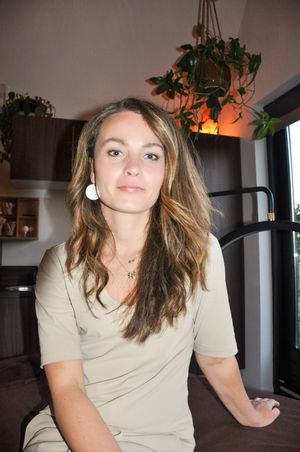 Young woman with long brown hair wearing a beige top sitting indoors with hanging plants in background.