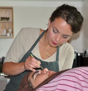 Esthetician with dark updo hair in gray apron performing precise facial treatment on a client.