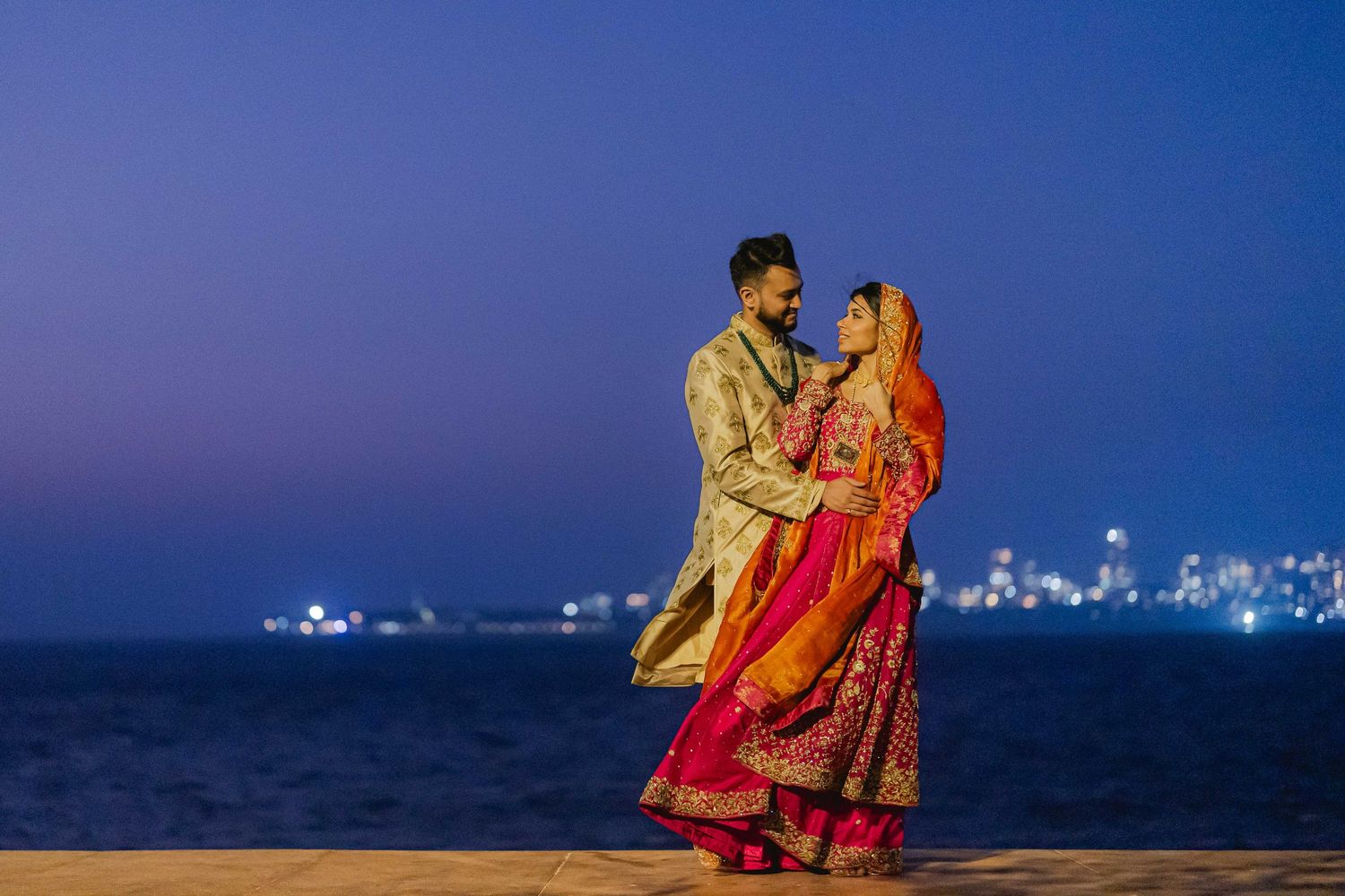 Indian couple walking together, bride in red saree and groom in black suit.