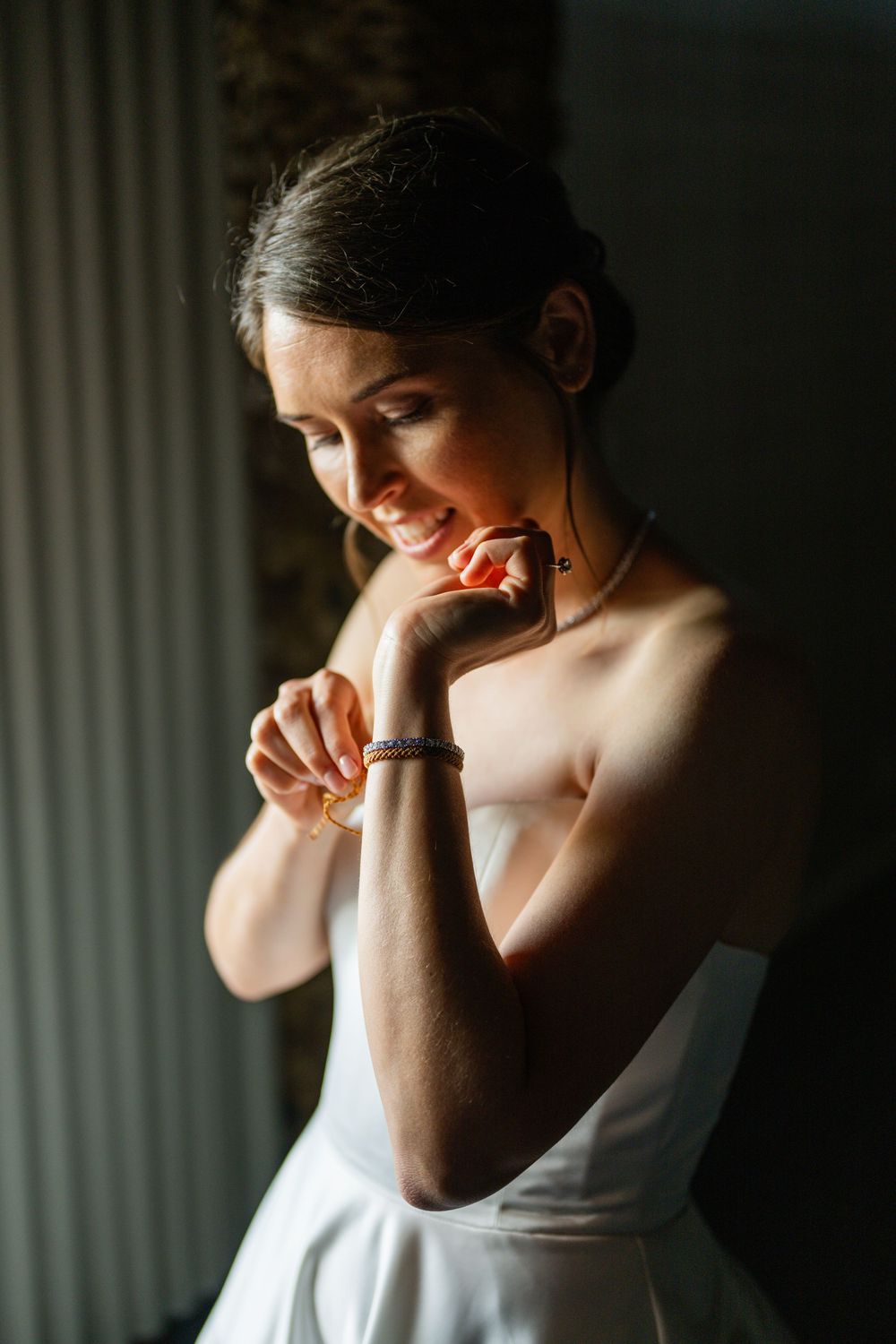 Smiling bride in strapless white gown adjusting bracelet in soft golden light, wearing pearl necklace and earrings.