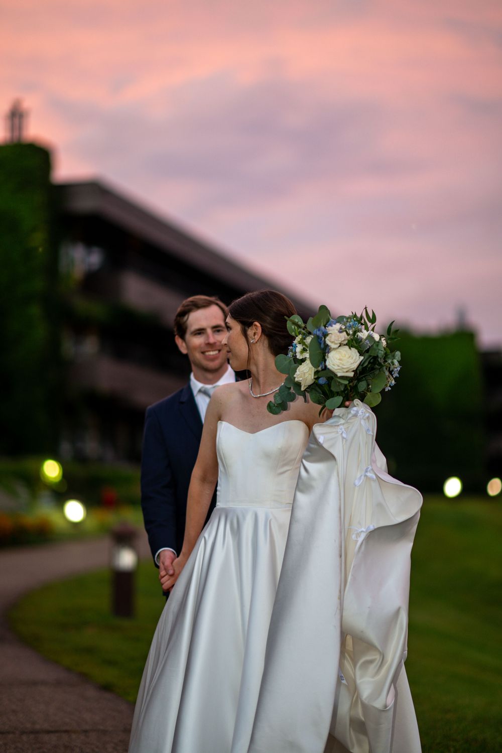 Bride and groom share a romantic moment at sunset, she holds a white bouquet while he smiles warmly in a navy suit.