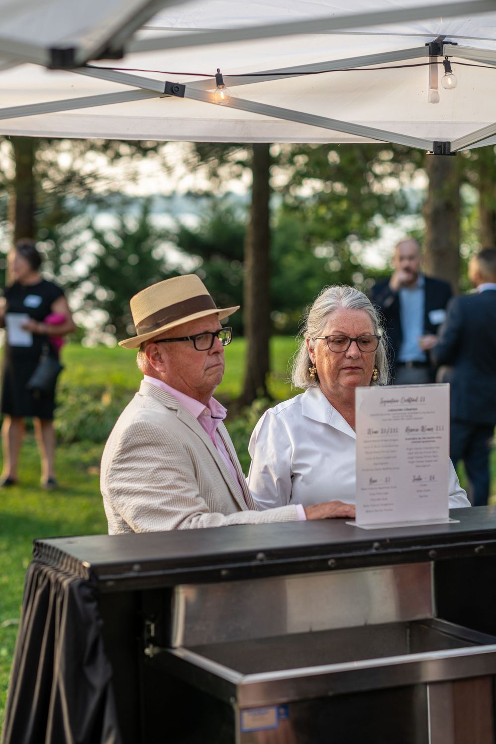 Couple reviewing a certificate at an outdoor evening event under a white canopy tent on a green lawn.