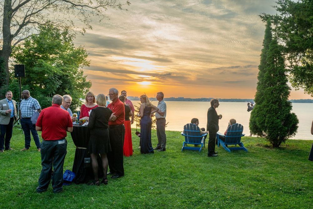 Group of people gathered on a lakeside lawn at sunset, socializing outdoors during a scenic evening event.