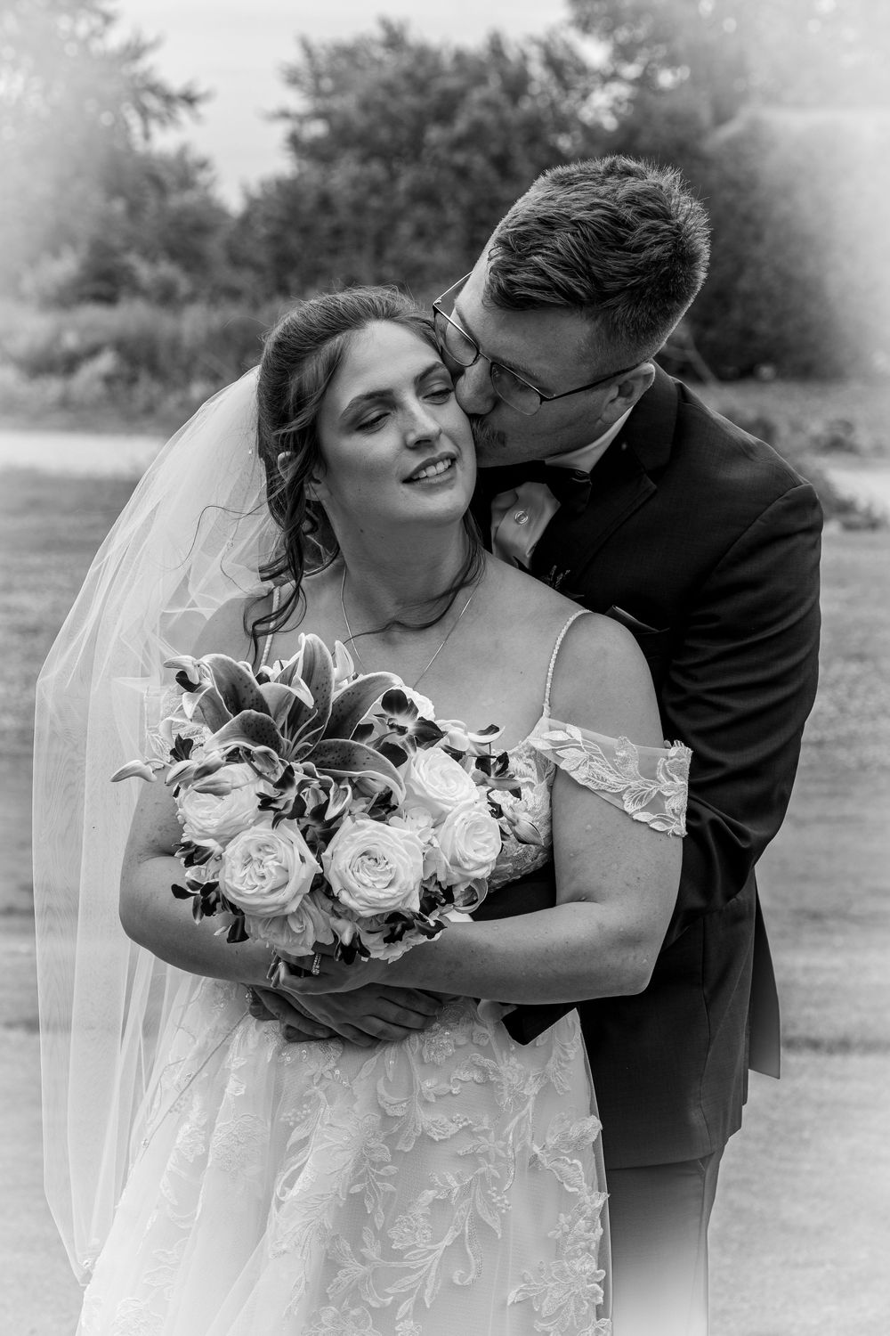 Black and white wedding photo of bride holding bouquet while groom kisses her cheek outdoors.