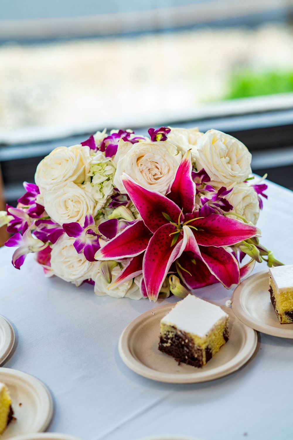 Elegant floral centerpiece with white roses, pink stargazer lilies, and purple orchids on a white tablecloth beside dessert plates.