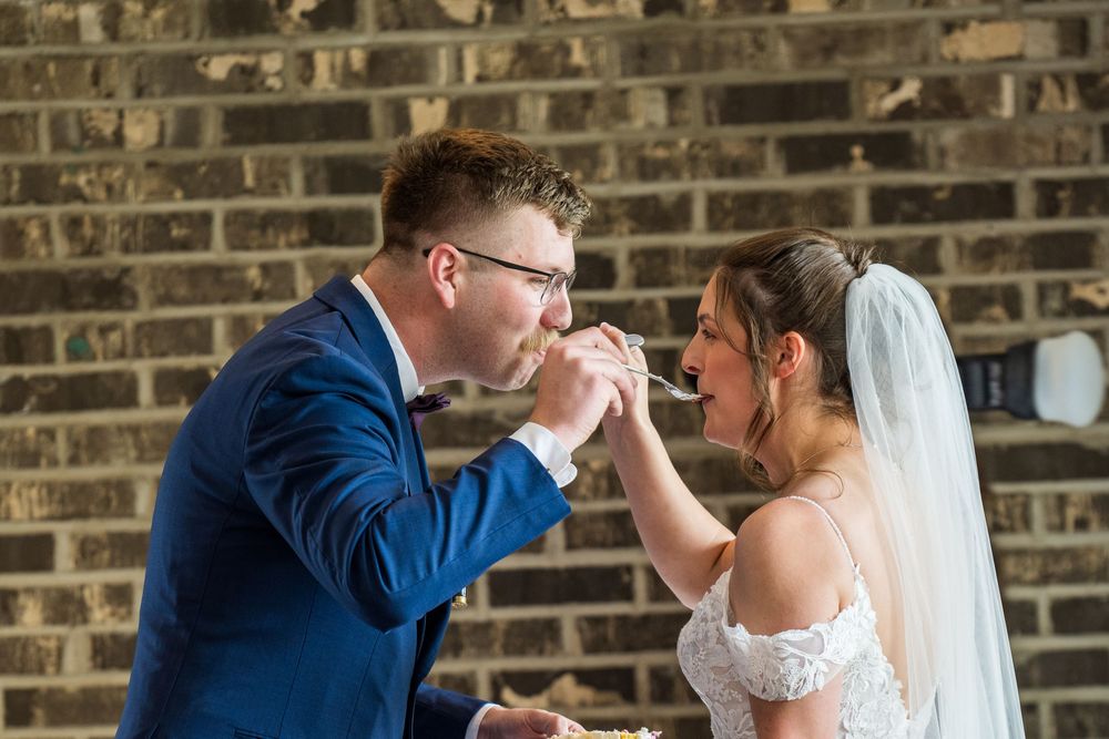Bride and groom sharing wedding cake at their reception near a brick wall.