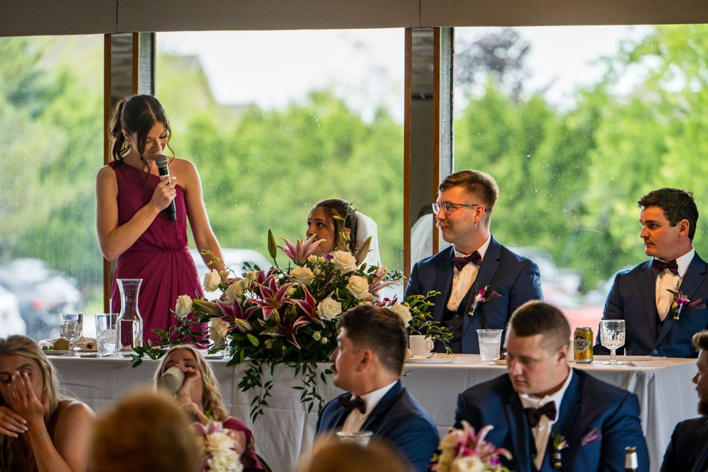 Bridesmaid giving a heartfelt wedding speech at a floral-decorated head table with bridal party seated nearby.