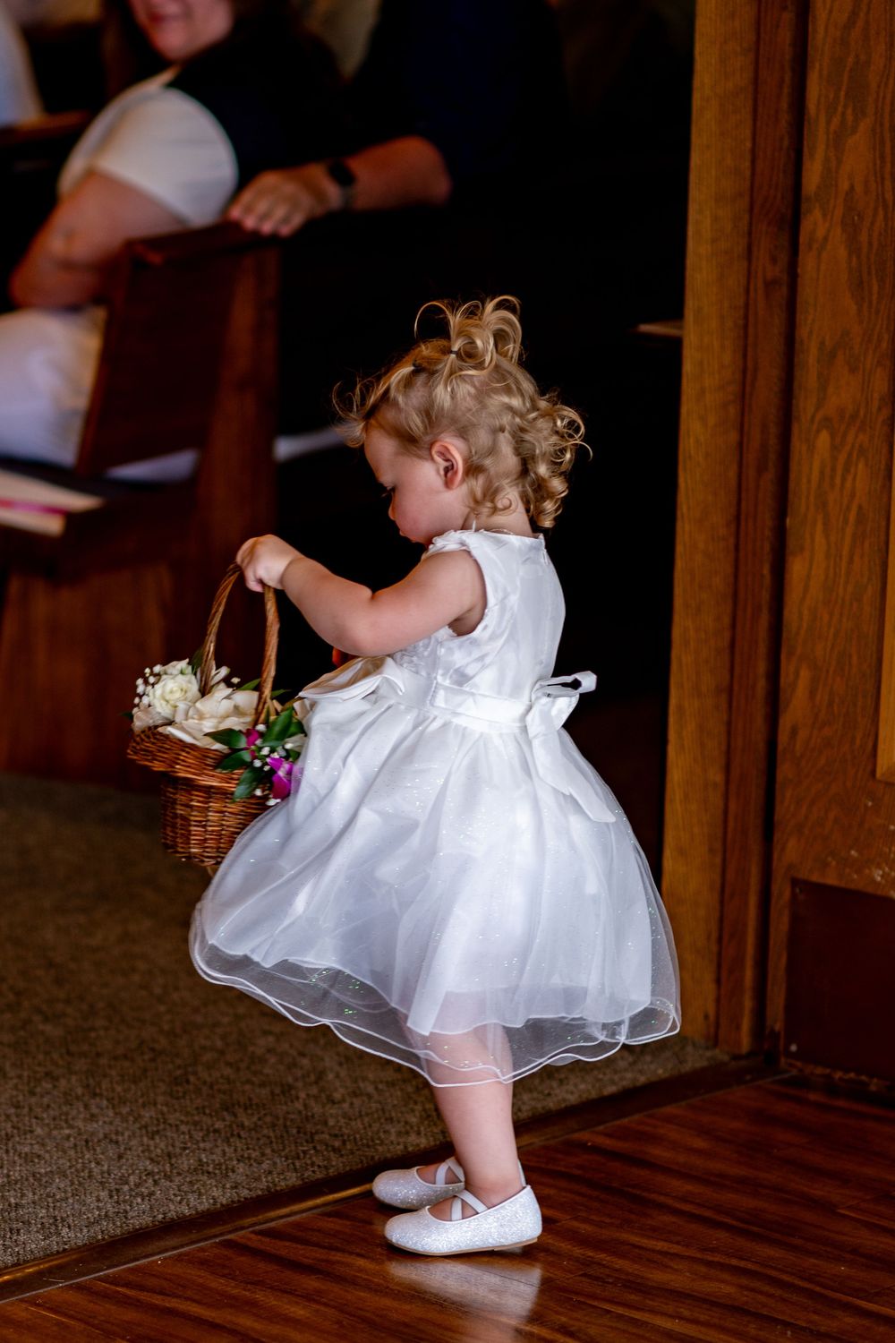Adorable flower girl in white dress carrying a wicker basket with flowers at a wedding ceremony.