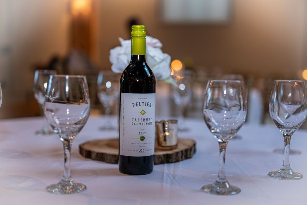 Peltier Cabernet Sauvignon wine bottle centerpiece on wood slice surrounded by crystal wine glasses on white tablecloth.