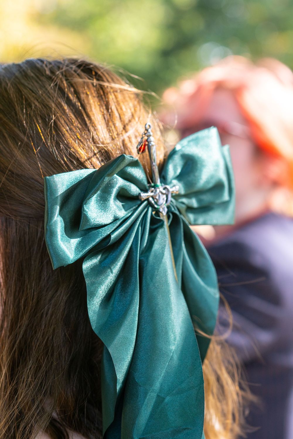 Close-up of a teal satin hair bow with a decorative brooch pin on a brunette girl's hair outdoors.