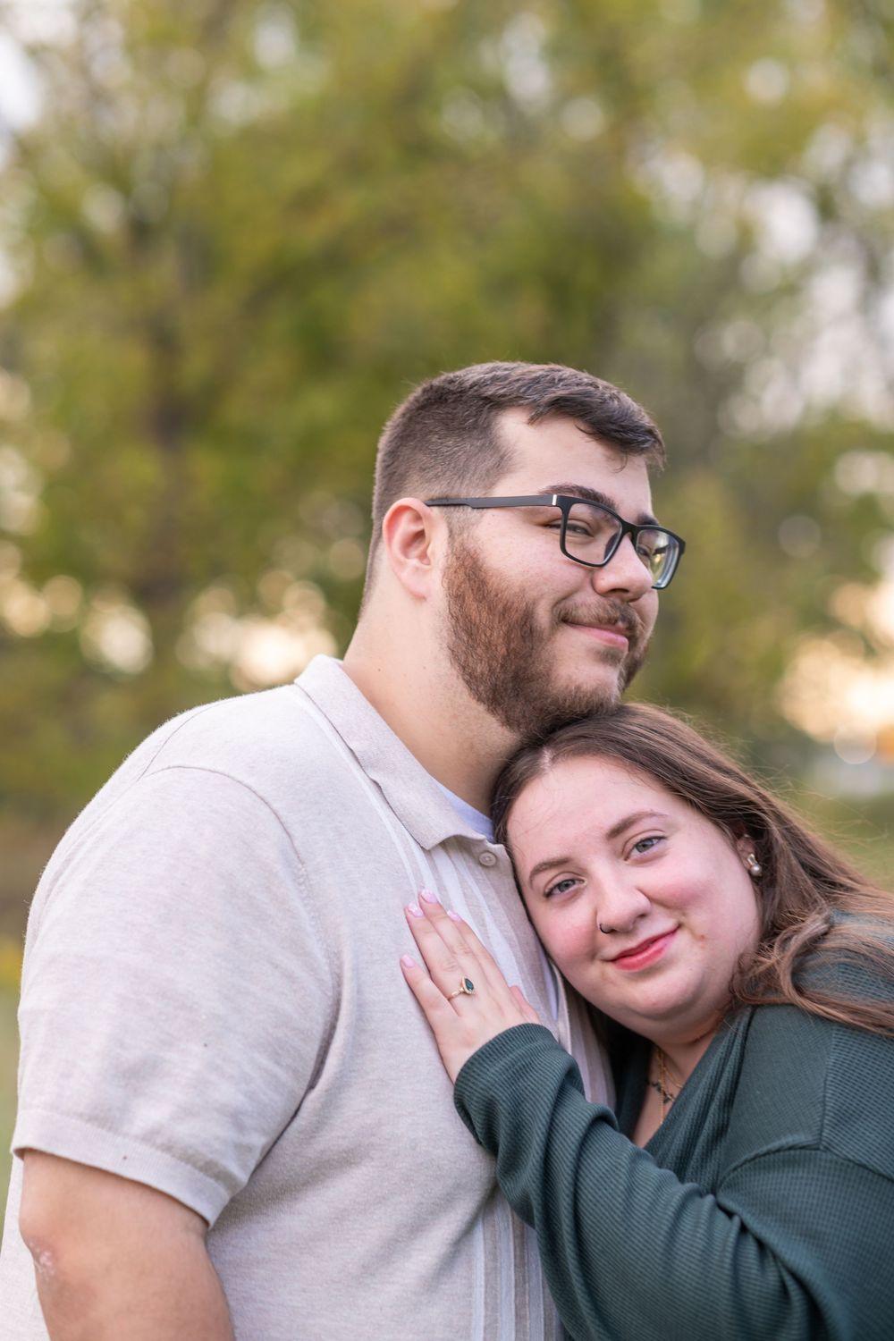 A couple embraces outdoors, woman in green smiling at camera, man in gray polo with glasses, bokeh tree background.