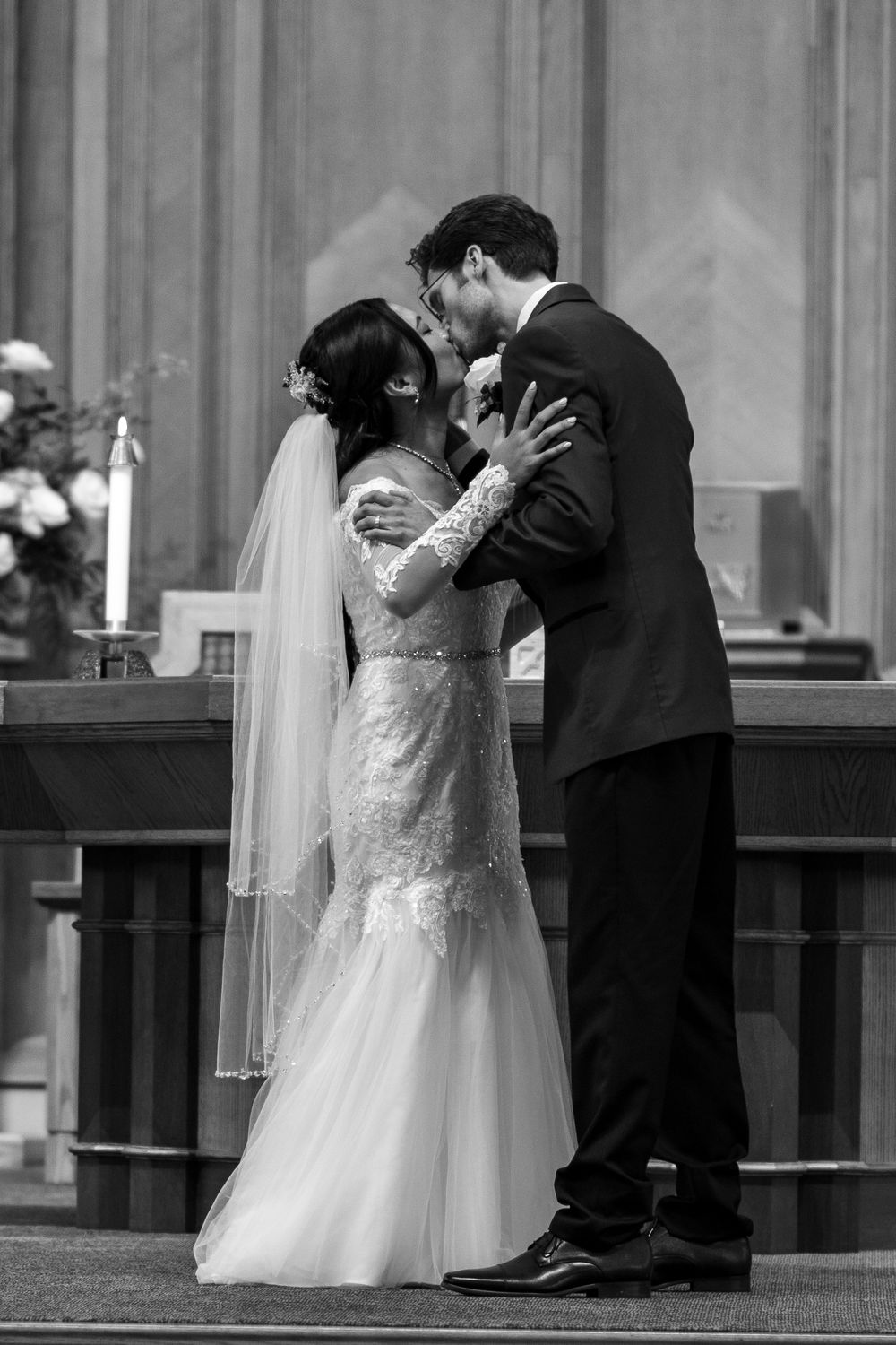 Black and white photo of bride and groom sharing first kiss at church altar during wedding ceremony.