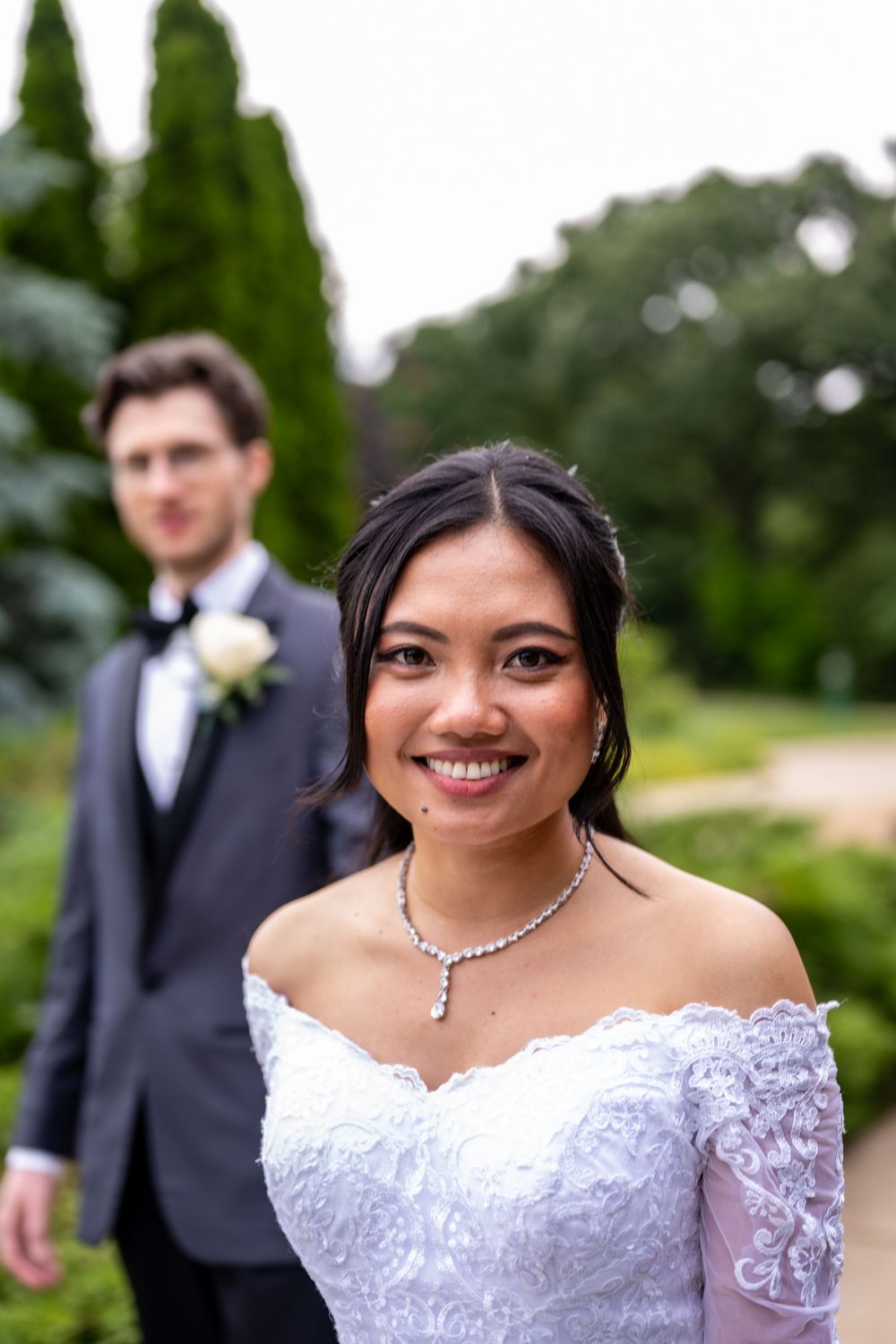 Smiling bride in white off-shoulder lace gown with diamond necklace, groom in dark suit with boutonniere in garden background.