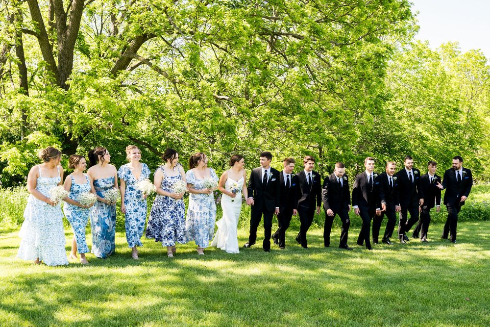 Wedding party walks outdoors on lush green lawn, bridesmaids in blue floral dresses and groomsmen in black suits.