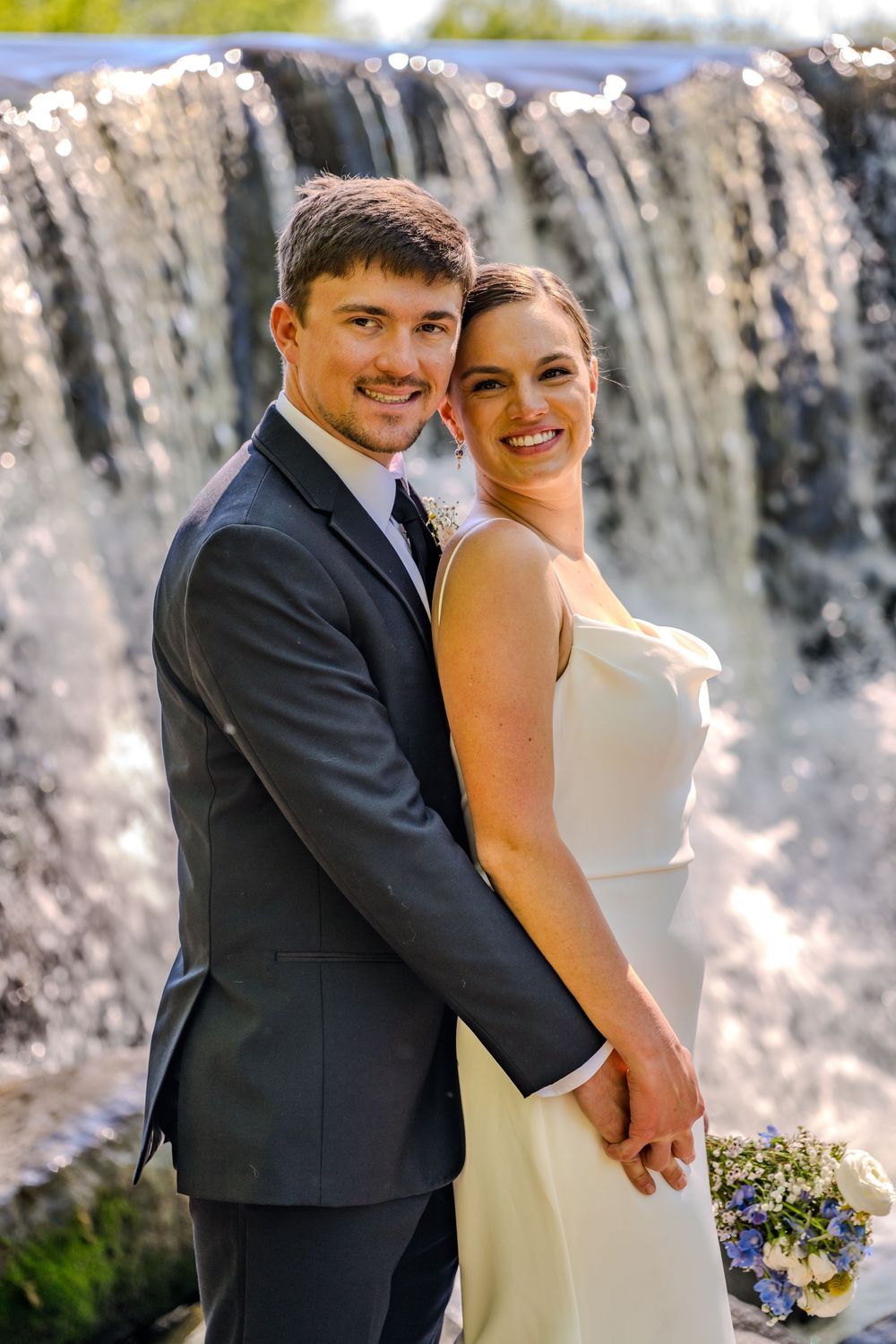 Smiling bride and groom pose together in front of a cascading waterfall, bride holding a blue and white bouquet.
