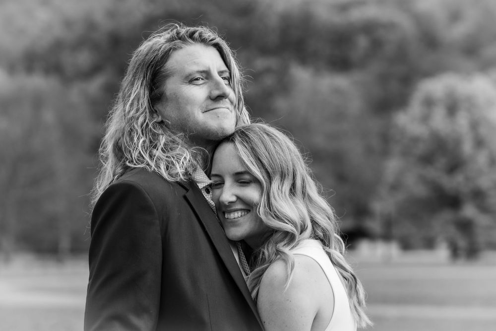Black and white photo of a couple embracing outdoors, woman smiling against mans chest, trees in background.