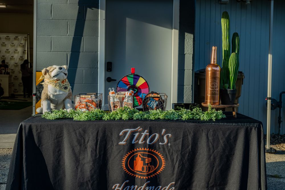 Tito's Handmade Vodka branded table display with decorations, a spin wheel, copper still, and cactus at an outdoor event.
