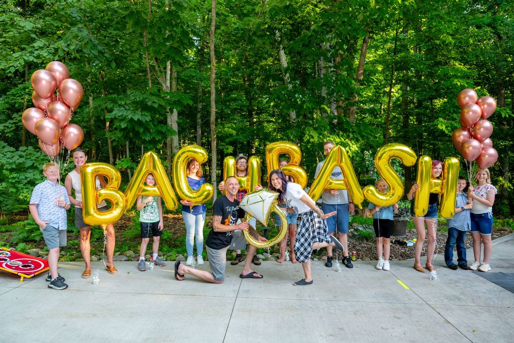 Group of people holding gold BACH BASH balloon letters outdoors with rose gold balloons and lush green trees in background.