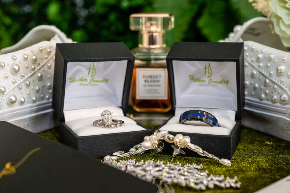 Two wedding rings in black boxes beside a perfume bottle and pearl accessories on a mossy surface.