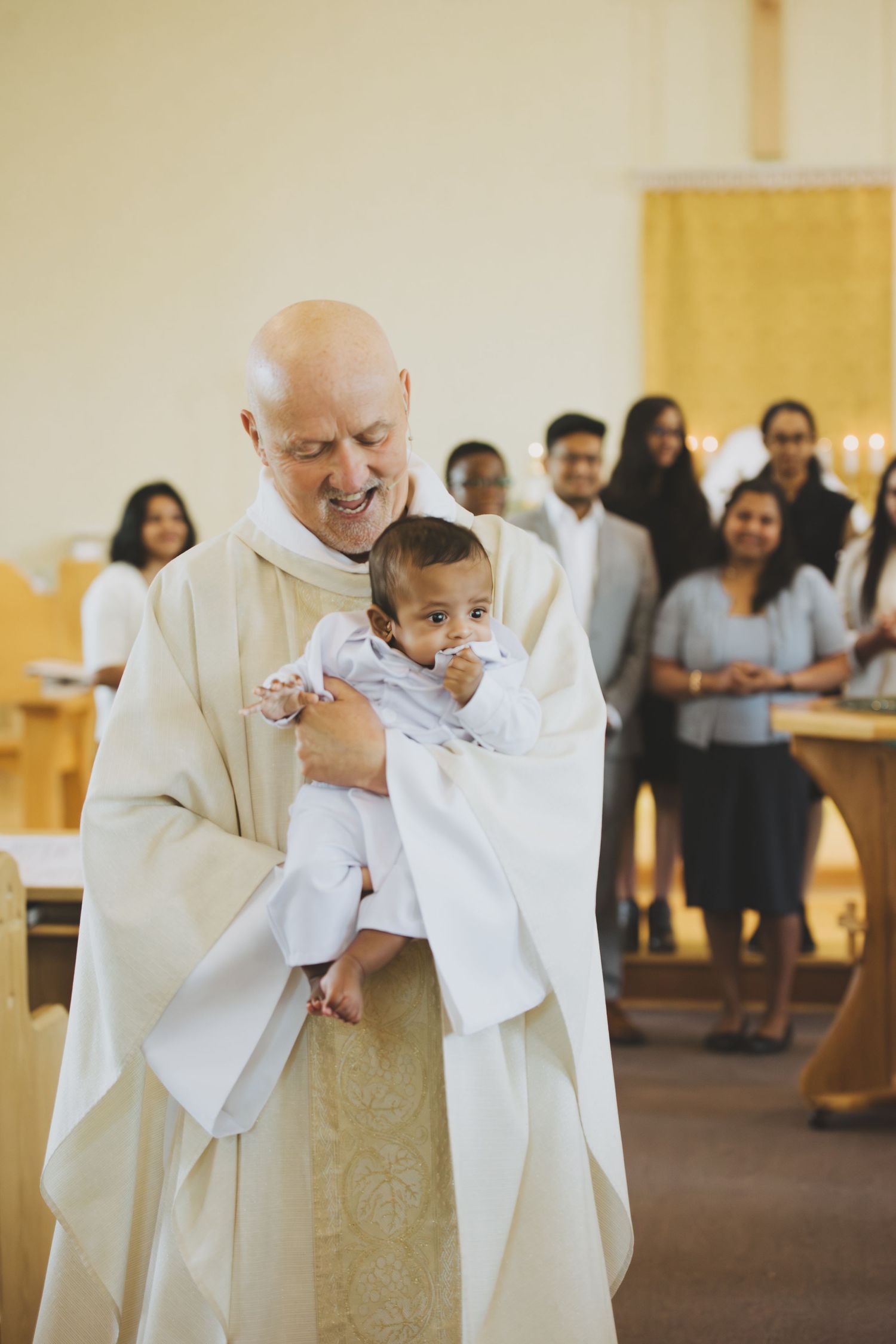 A smiling priest in white vestments holds a baby in white baptismal gown during a church christening ceremony.