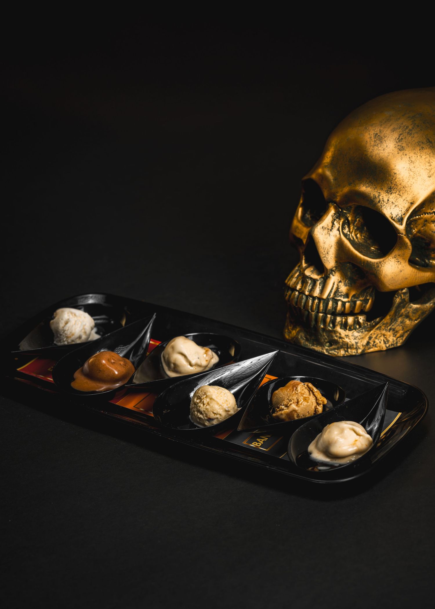Elegant black tray with assorted ice cream scoops and sauces in spoon-shaped dishes on dark background.