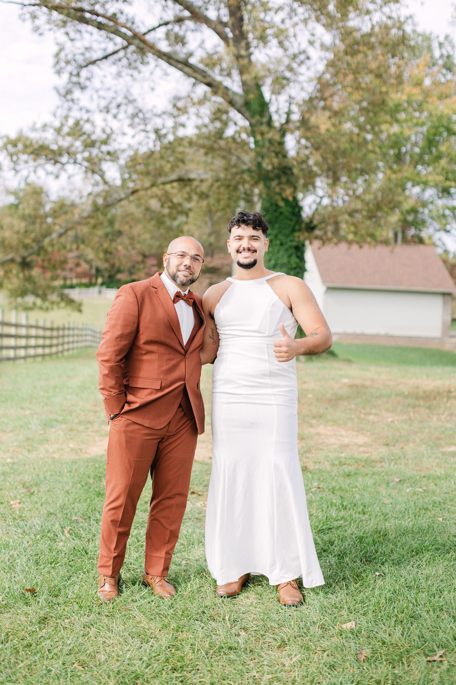 Two grooms pose outdoors on green grass, one in a rust suit and one in a white gown, at a rustic farm wedding.