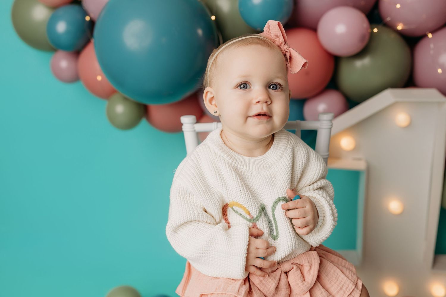 Baby girl in white sweater sits before teal backdrop with colorful balloons at her first birthday celebration.