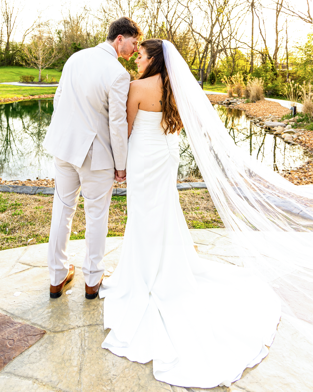 Bride and groom hold hands facing a serene pond at sunset, brides cathedral veil trailing behind.