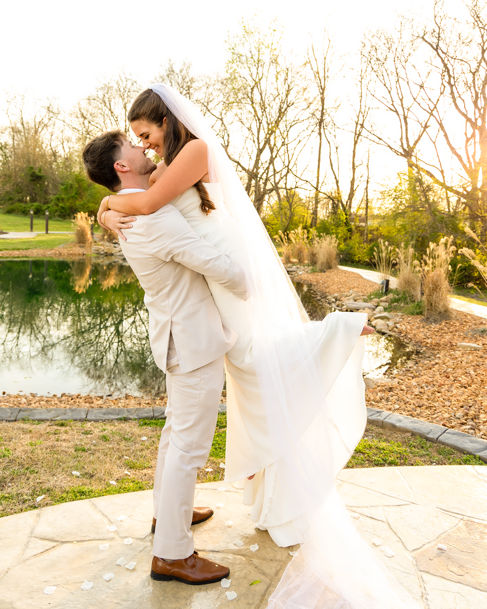 Groom lifts bride in white gown and veil for romantic kiss by pond at golden hour wedding.