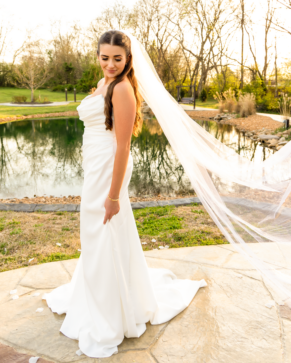Elegant bride in strapless white gown with flowing cathedral veil poses by reflective pond.