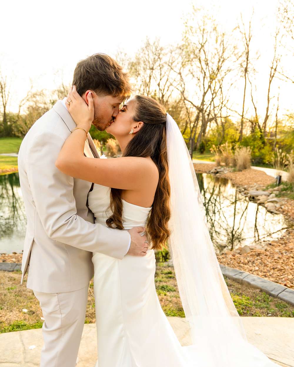 Bride and groom share passionate kiss in beige suit and white wedding dress by pond.