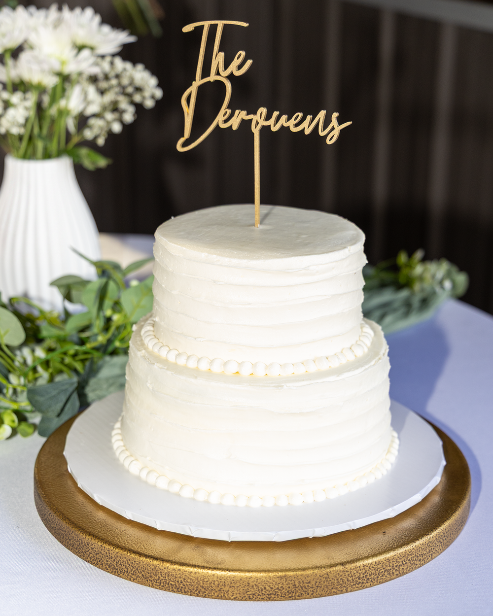 Two-tier white wedding cake with gold The Deroyens cake topper on gold stand.