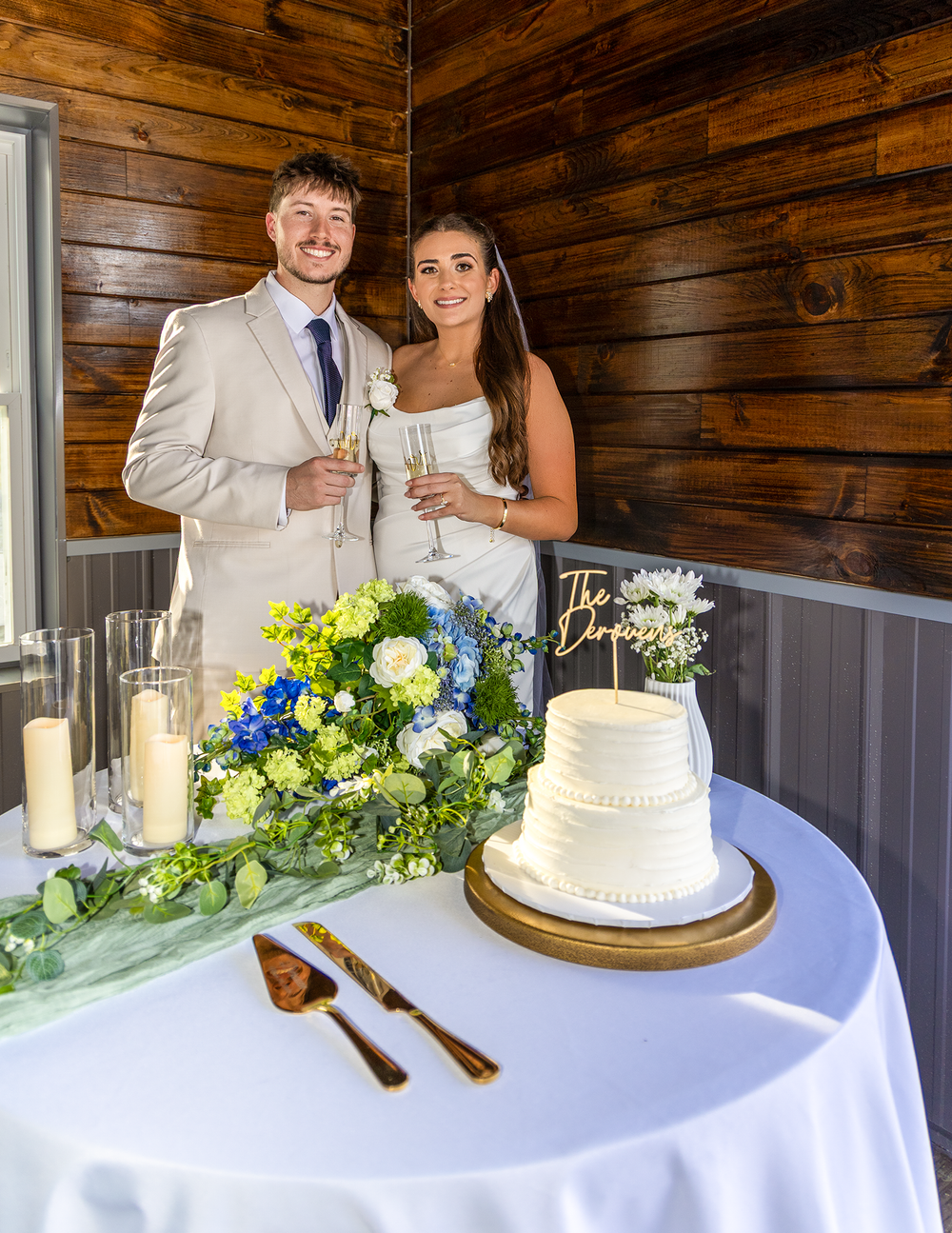 Newlywed couple smiles beside two-tier wedding cake with champagne glasses in rustic venue.