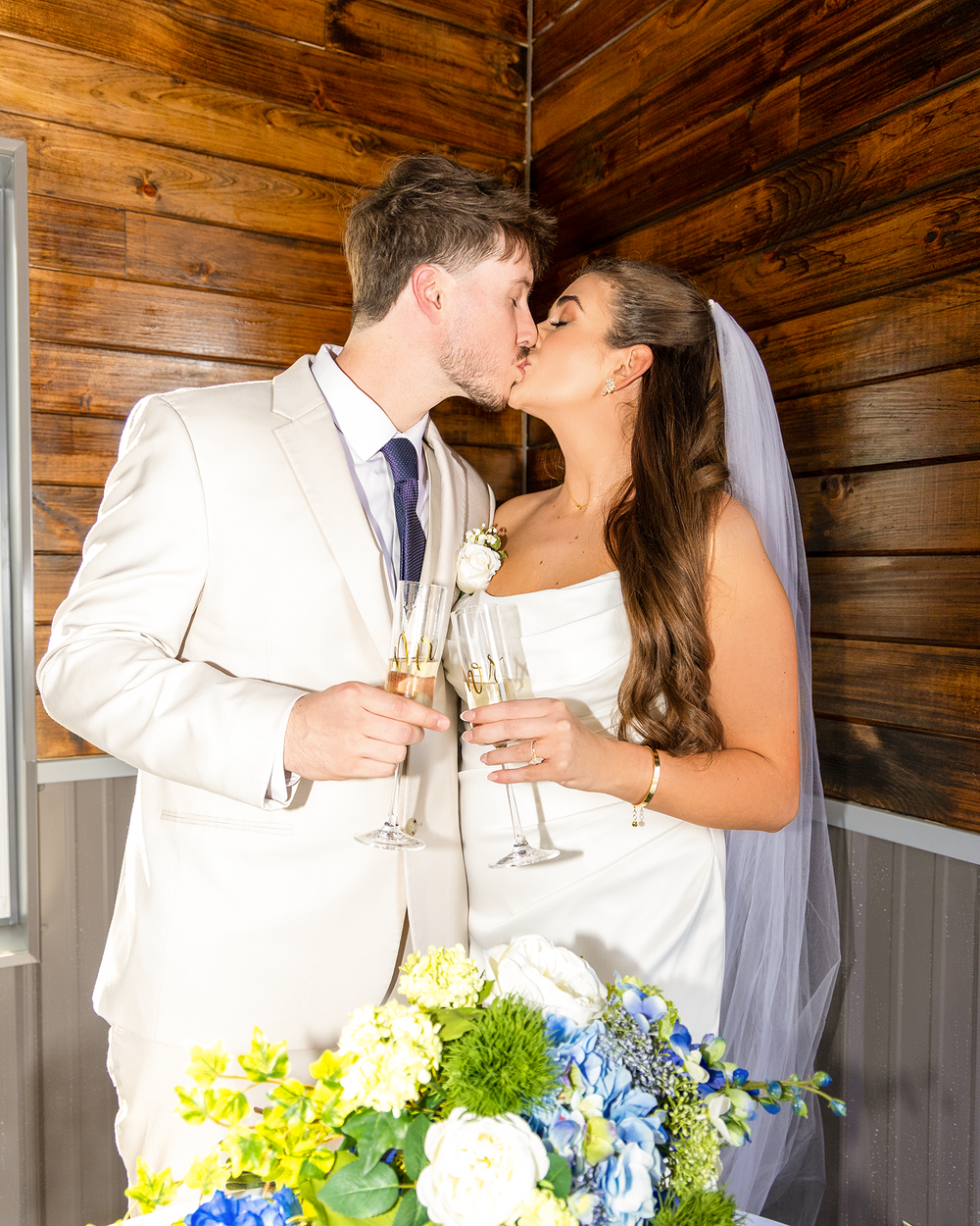 Bride and groom share a champagne toast kiss in front of rustic wood wall with blue and white floral arrangement.