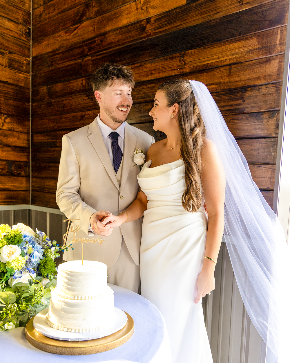 Smiling bride in white gown and groom in tan suit stand together at wedding cake table in rustic venue.