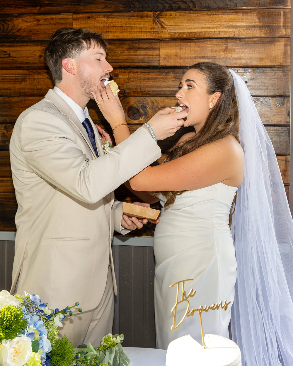 Bride and groom playfully feed each other wedding cake at rustic barn reception venue.