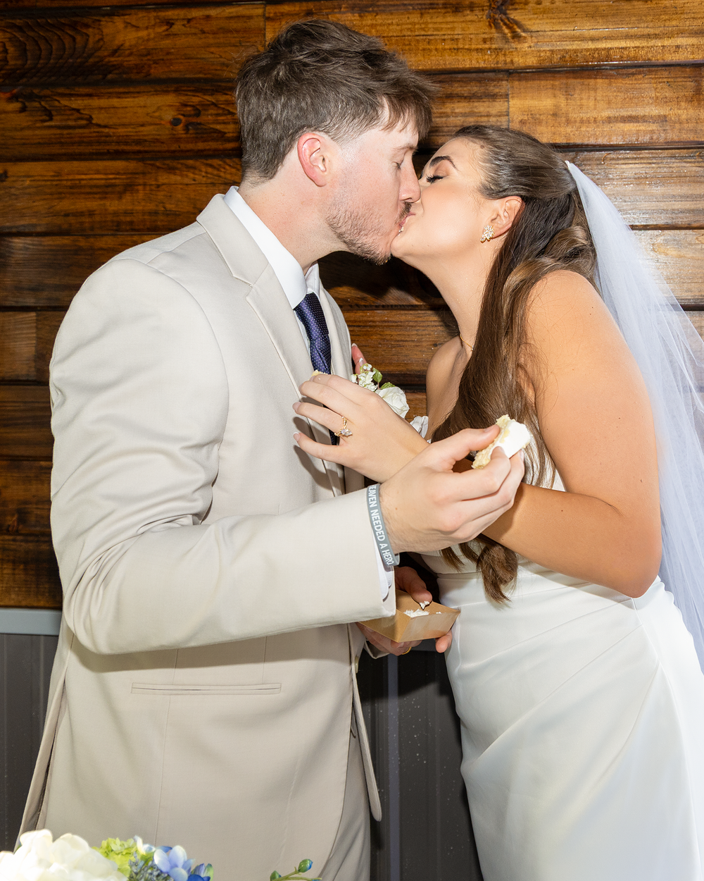 Newlyweds share a romantic kiss while holding wedding cake at their rustic barn wedding reception.