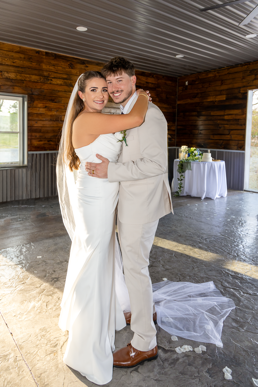 Bride in white gown and groom in tan suit embrace and smile inside rustic wood-paneled wedding venue.