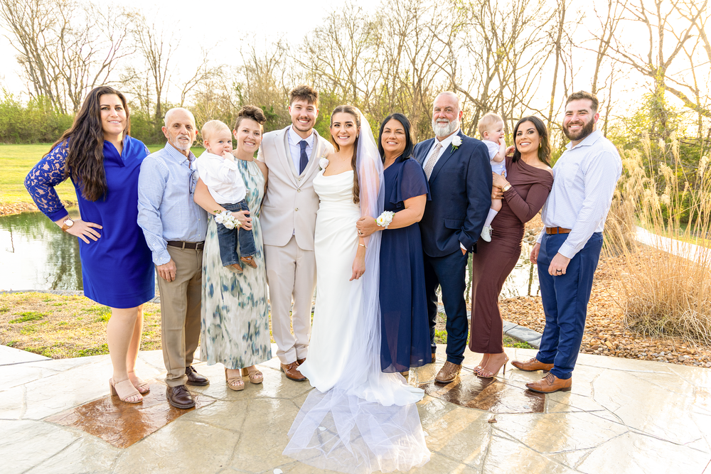 Wedding party family portrait of eleven guests posing outdoors at golden hour near a pond.