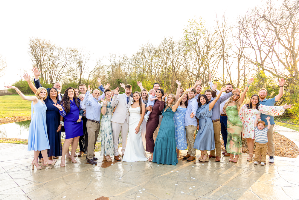 Large group of joyful wedding guests celebrating outdoors near a pond at golden hour.