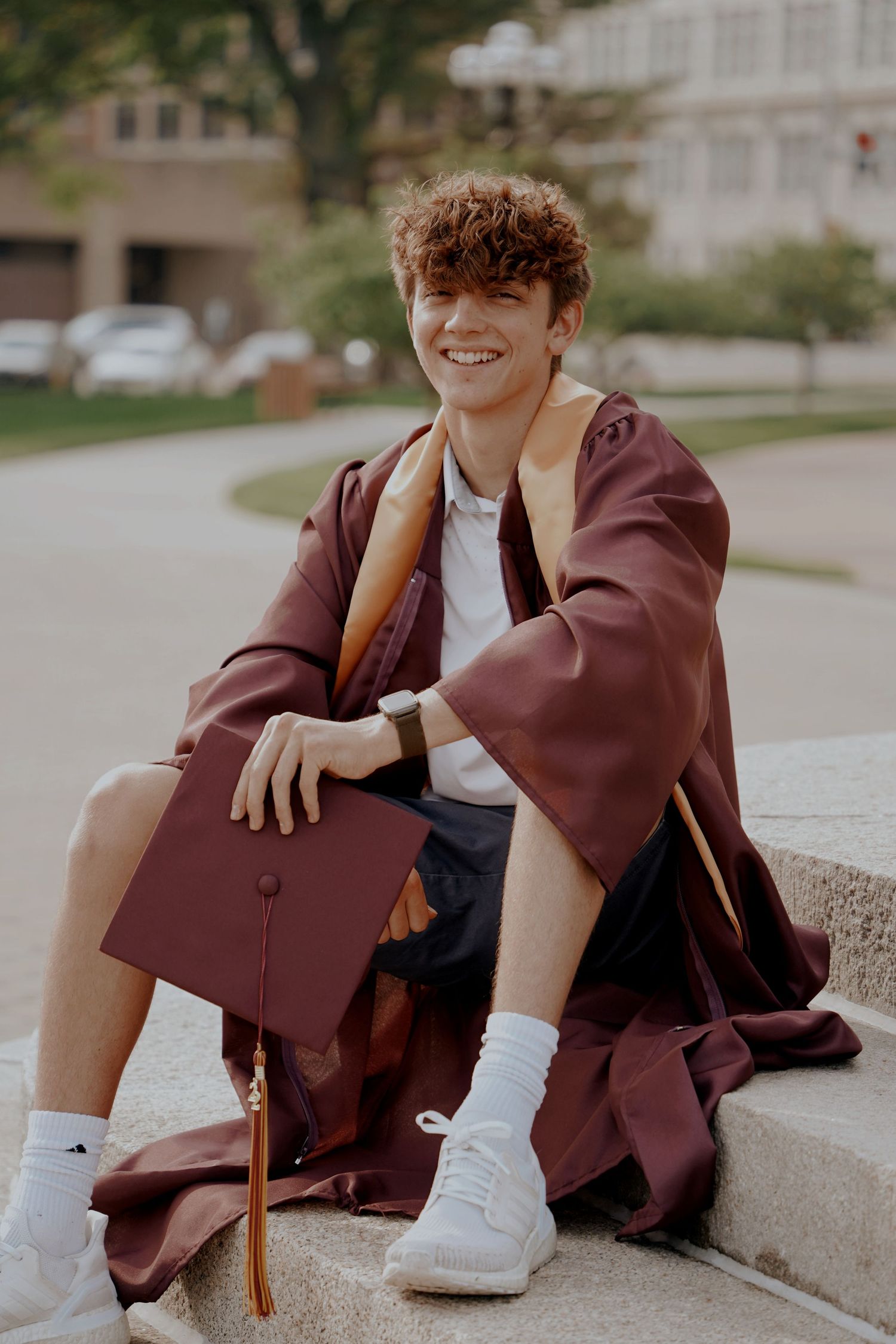 Smiling young man in maroon graduation gown and cap sitting on outdoor steps.