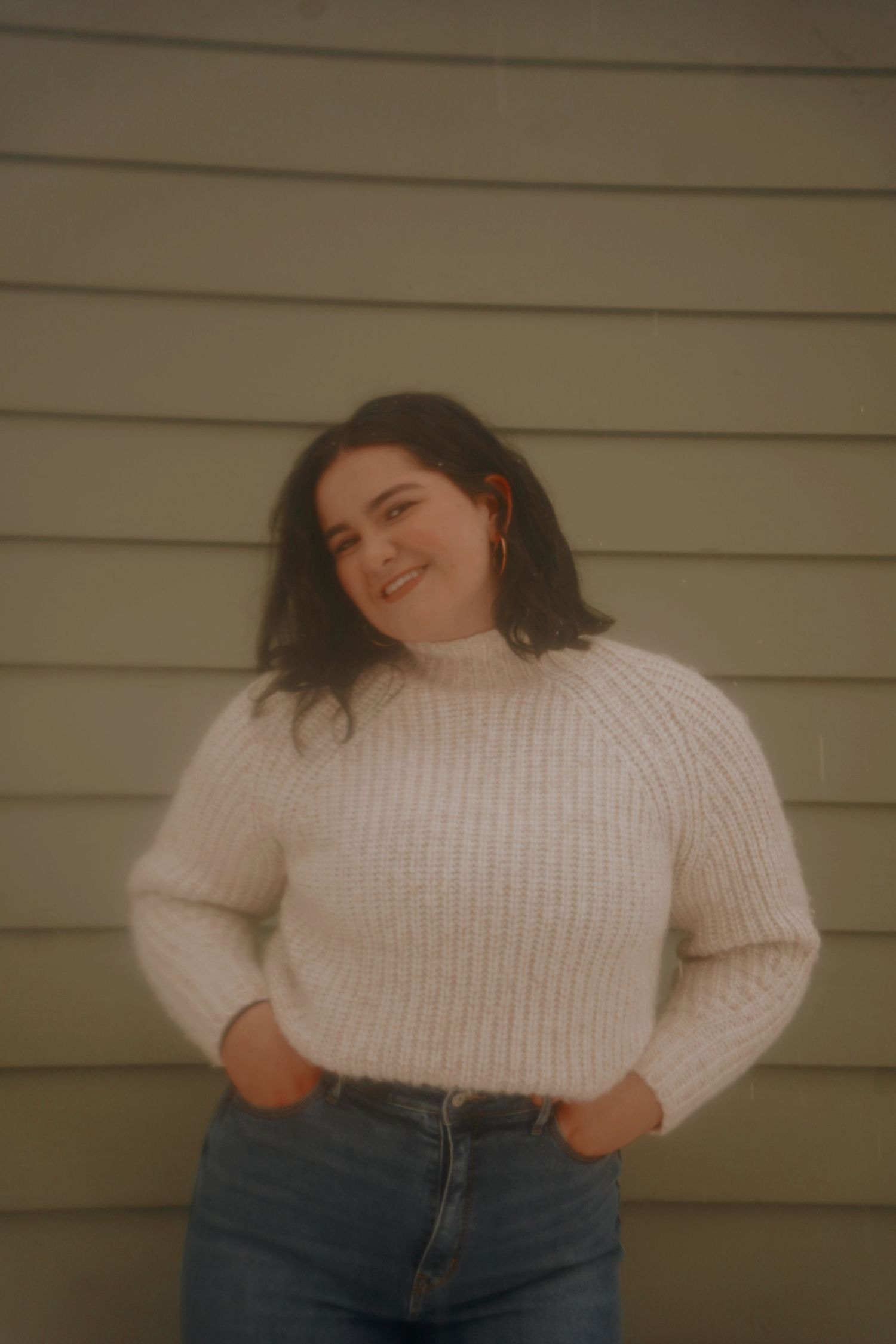 Young woman in cream ribbed turtleneck sweater and jeans smiling against a gray wooden wall.