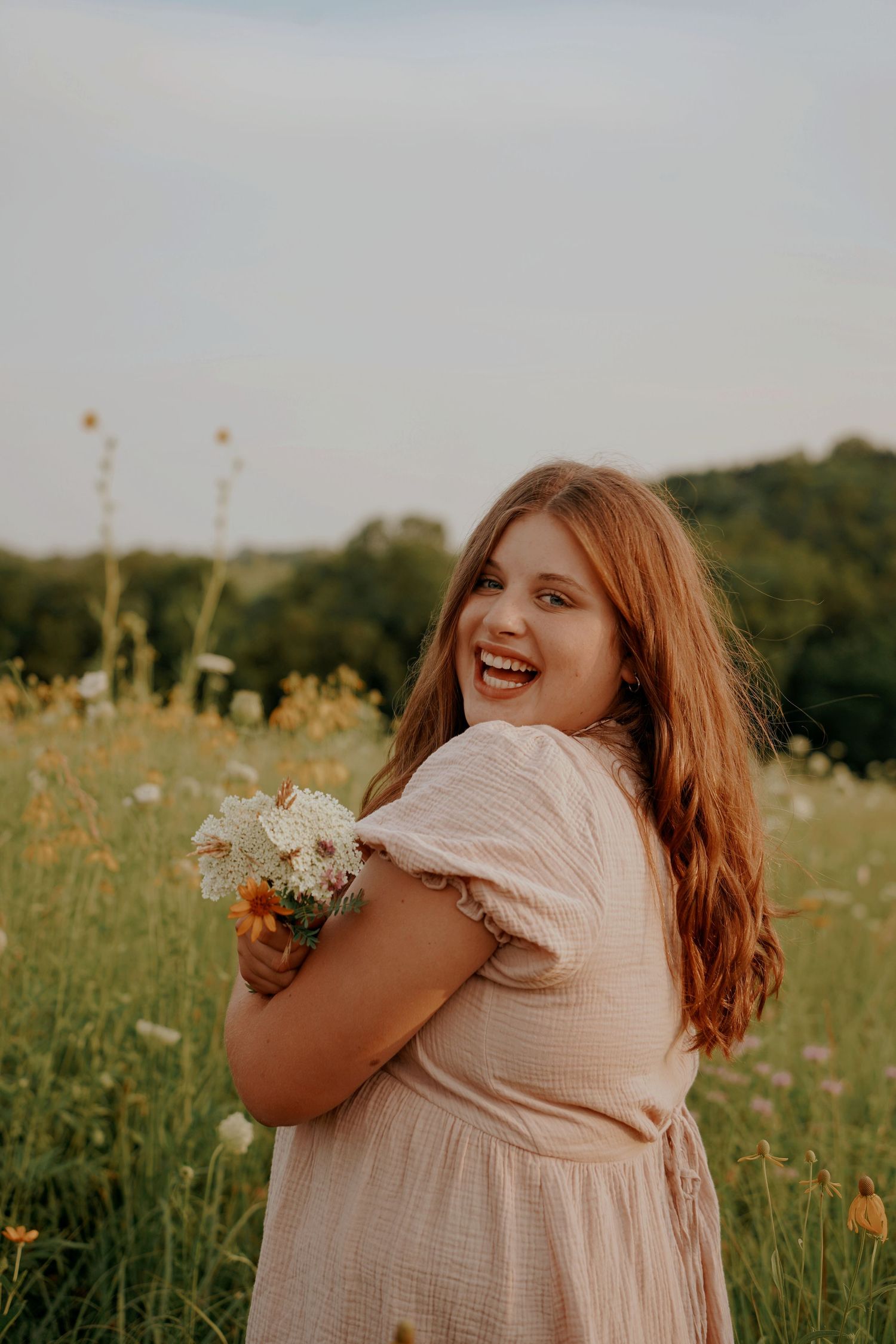 Joyful young woman in pink dress holding wildflowers while laughing in a sunny meadow.