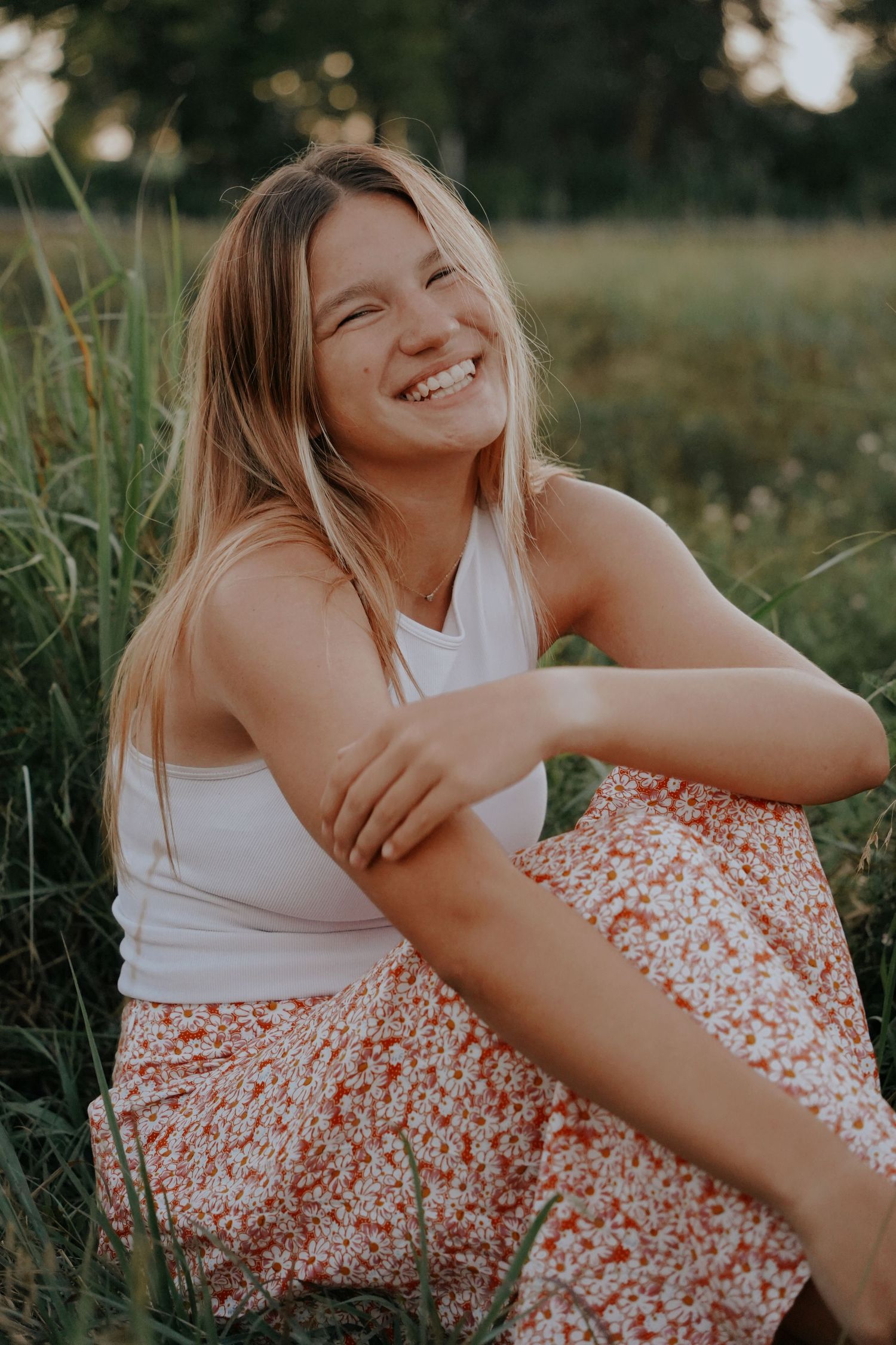 Smiling young woman in white tank top and floral skirt sitting in a lush green meadow.