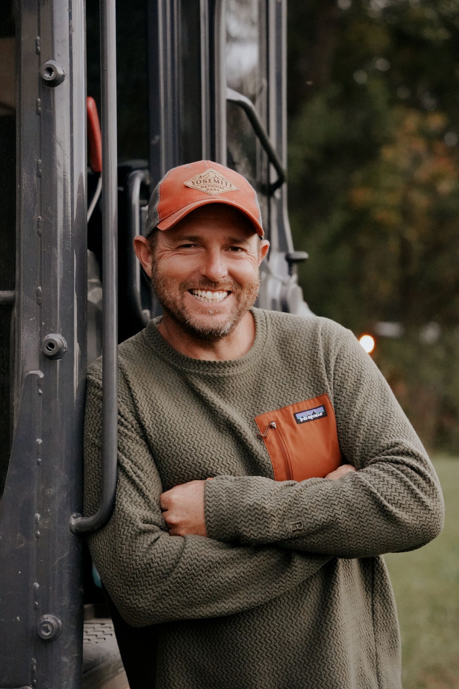 A smiling man in an orange cap and olive sweater leans against a vintage vehicle outdoors.