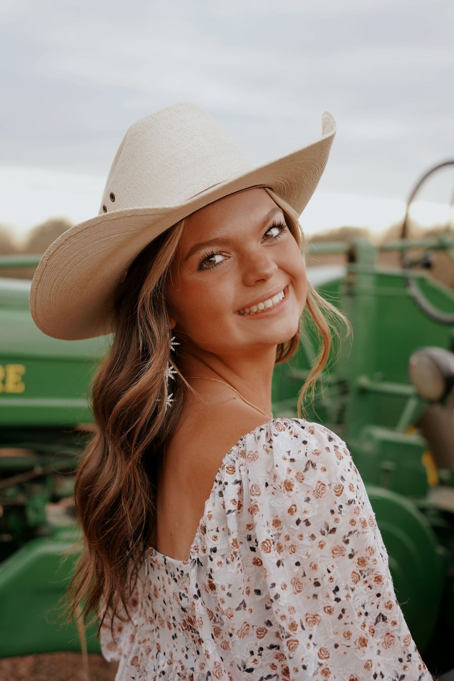Smiling young woman in a cream cowboy hat and floral top poses near green farm equipment outdoors.