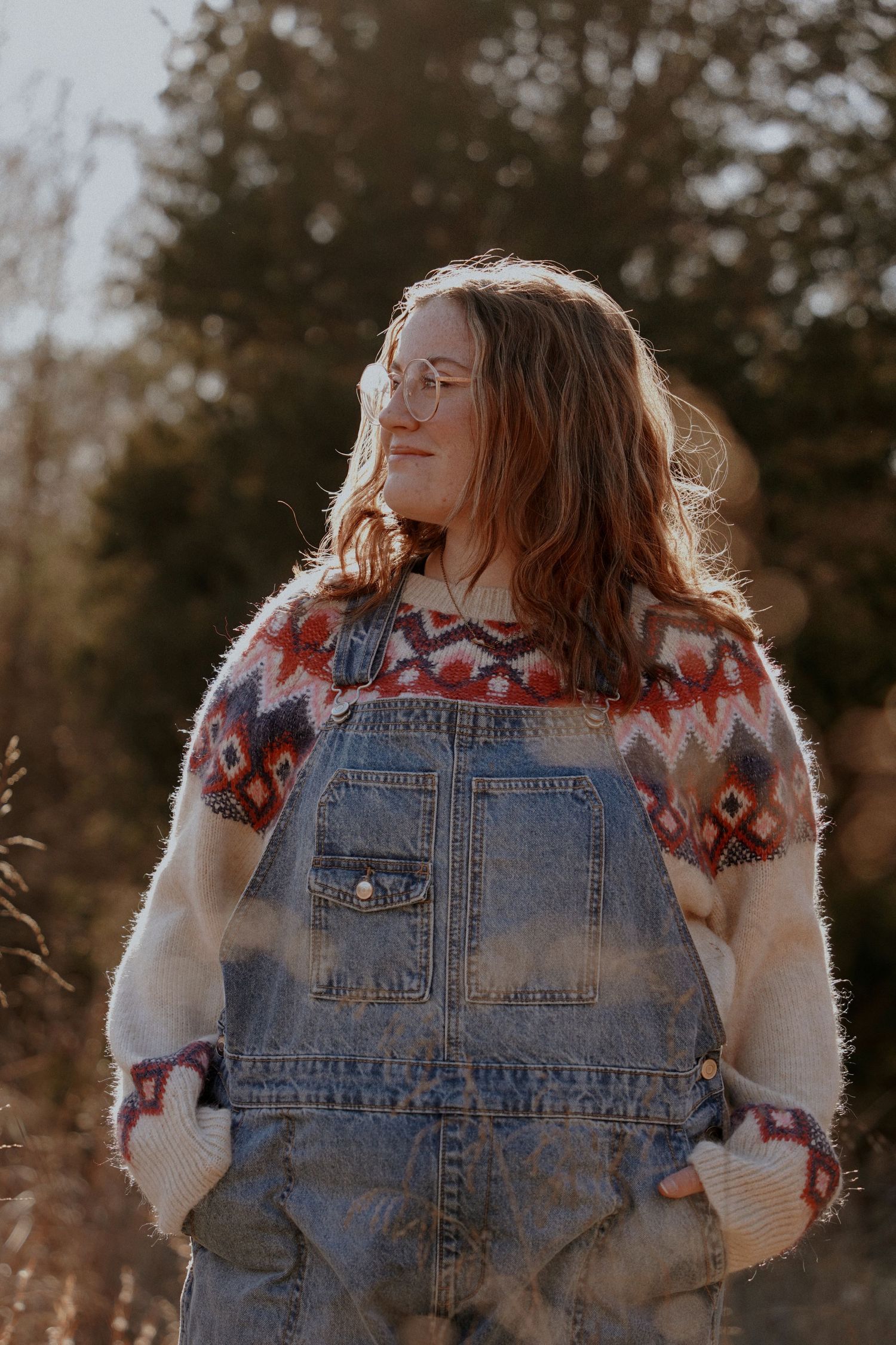 Young woman in denim overalls and patterned sweater stands in golden sunlight among autumn trees.