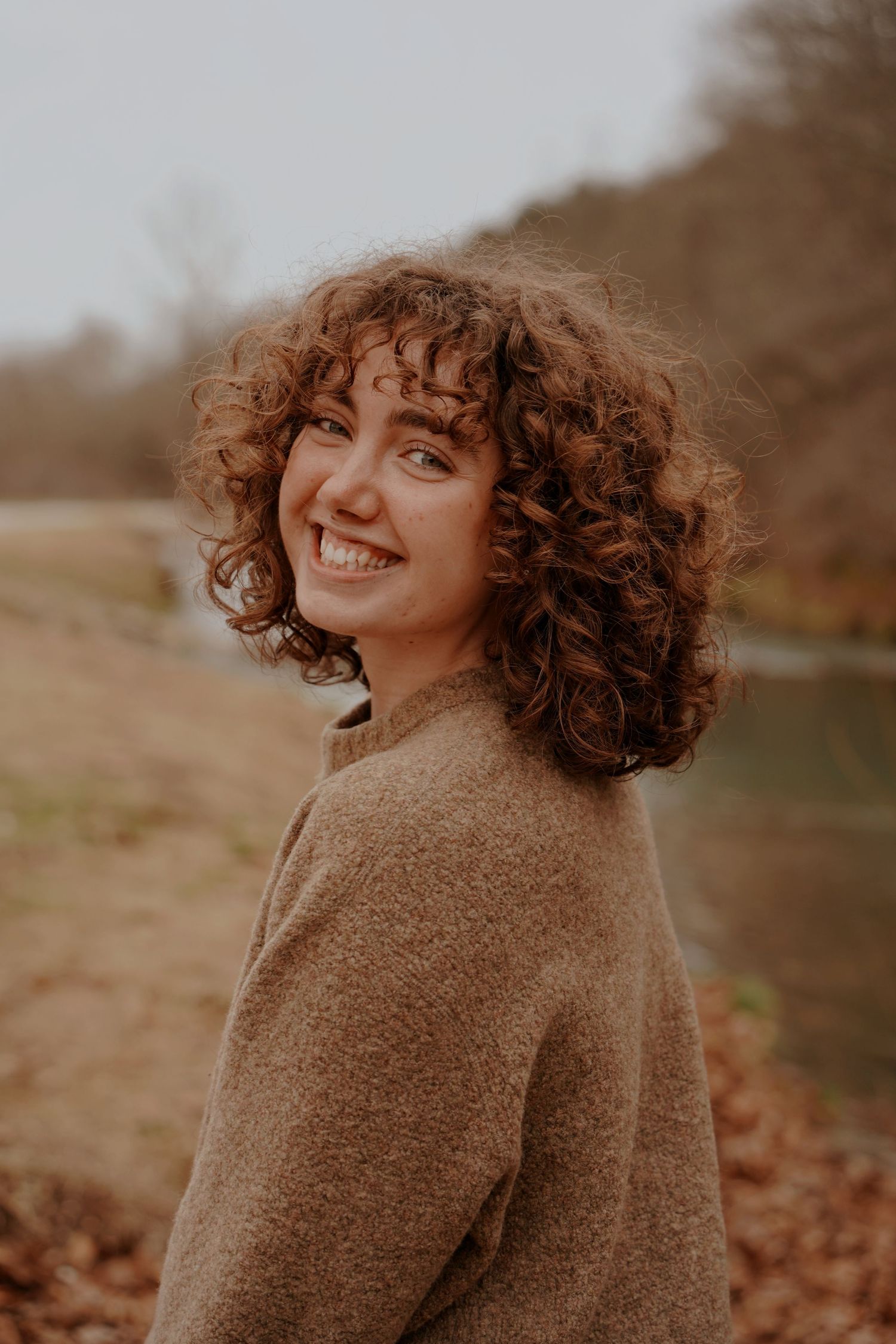 Curly-haired young woman in a brown sweater smiles over her shoulder near a reflective outdoor pond.